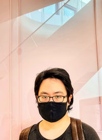 Portrait of young man wearing eyeglasses and face mask against reflective glass background.
