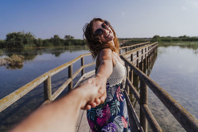 Woman holding hand of boyfriend while standing on footbridge against sky