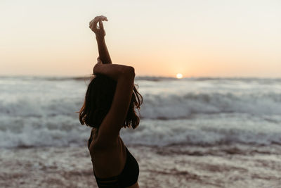 Side view of unrecognizable female in bikini raising hands up and contemplating seascape in sunset in summertime