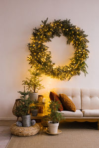 Christmas trees in pots near a cozy sofa over which a wreath with lights hangs on the wall