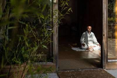 Full body front view of focused bearded ethnic senior male practicing chi kung meditation in lotus position in dark studio