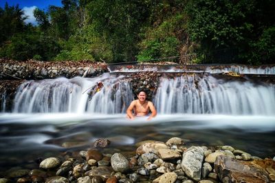 Portrait of shirtless man sitting amidst waterfall in forest