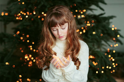 Happy young girl sitting by the christmas tree with garland