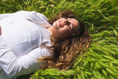 From above of optimistic female lying on green grass with closed eyes while resting on sunny summer day in nature