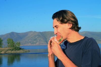 Close-up of man eating burger while standing at lakeshore against blue sky