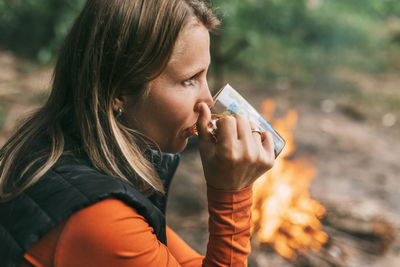 Close-up of an adult woman sitting with a cup of tea by the fire while relaxing in the forest