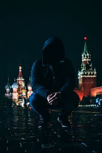 Young man in hooded shirt crouching on wet street at night