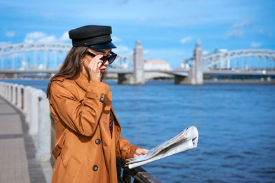 Stylish young woman reading fresh newspaper on embankment of river on sunny day