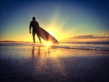 Silhouette surfer with surfboard walking on beach against sky during sunset