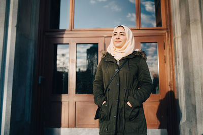 Confident young muslim woman wearing hijab standing with hands in pockets against door