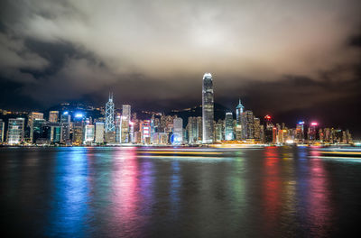 Illuminated city at waterfront against cloudy sky