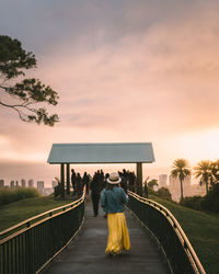 Rear view of people standing by railing against sky during sunset