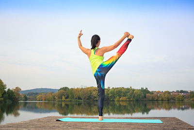 Rear view of woman exercising on pier over river against sky