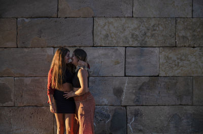 Two girls standing against stone wall during sunset