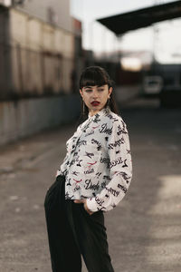 Pensive young ethnic lady with long dark hair in casual dress standing in street near old houses and looking away on sunny day