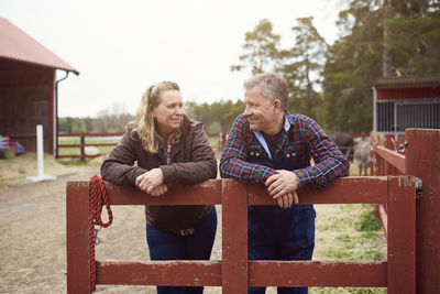 Smiling mature couple looking at each other while leaning on fence in farm