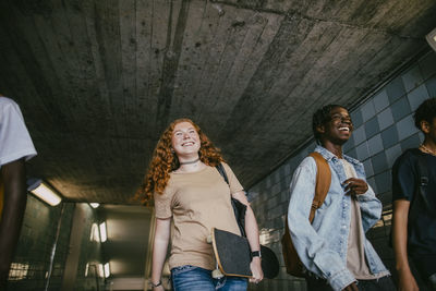 Low angle view of smiling teenage girl carrying skateboard while walking with friends in tunnel