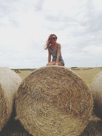 Smiling woman looking away while sitting on hay bales against sky