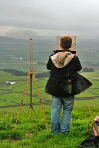 Full length rear view of painter standing on grassy hill against cloudy sky