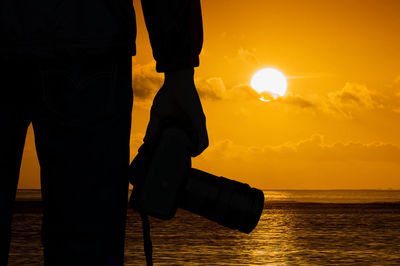 Close-up of silhouette man holding camera by sea during sunset