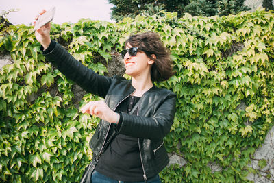 Smiling woman taking selfie with smart phone while standing against plants at park