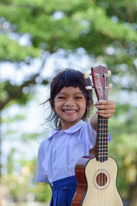 Portrait of smiling girl holding ukulele while standing against trees