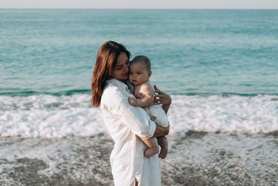 A young mother walks along the beach with a small child in diapers.