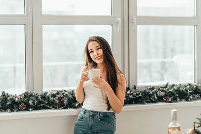 Portrait of smiling woman holding coffee cup against window