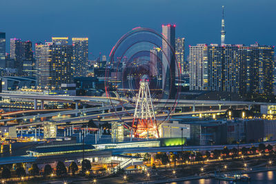 Illuminated ferris wheel by river and buildings against sky at night