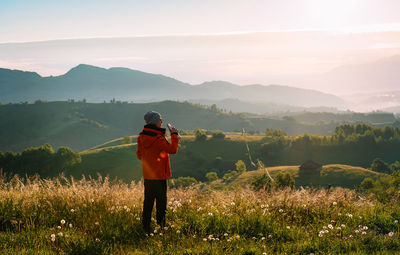 Rear view of man standing on field against mountains and drinking coffee in a summer morning