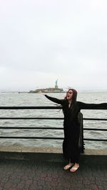 Happy young woman with arms outstretched standing by sea against clear sky