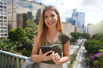Portrait of young brazilian woman holding mobile phone looks at camera, sao paulo, brazil