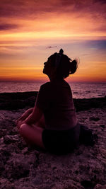 Young woman sitting on shore at beach against sky during sunset