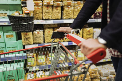 Midsection of man holding smart phone while pushing shopping cart in store