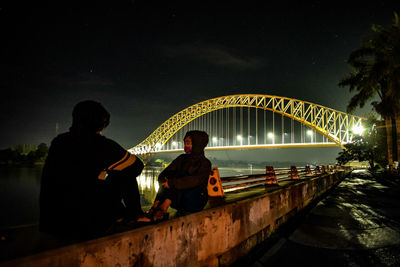 Friends sitting on retaining wall against sky at night