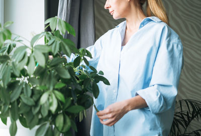 Young woman in blue shirt with spray with water in hands takes care of houseplant at home