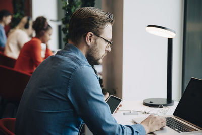 Side view of businessman using laptop while sitting at desk by colleagues in office