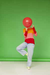 Woman covering face with red balloon standing on one leg against green background