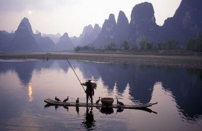 Man sailing boat with birds in river against mountains