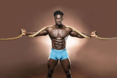 Portrait of shirtless athlete exercising with rope against brown background