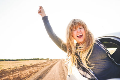 Young woman with arms outstretched leaning out from car window against clear sky