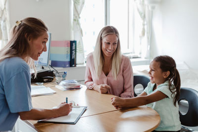 Female pediatrician and mother looking at smiling girl sitting at desk in hospital