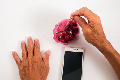 Close-up of man inserting money in piggy bank with mobile phone against white background