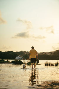 Full body of anonymous male owner with boots in hands walking in water near running dog on summer day in nature