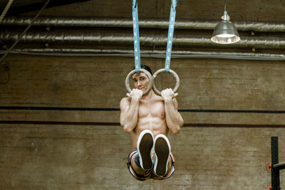Shirtless man hanging with gymnastic rings against wall in gym