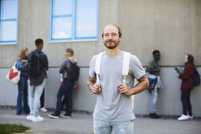 Portrait of smiling student carrying backpack standing at university campus with friends in background