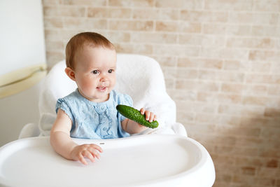 Cute baby girl holding cucumber while sitting on high chair at home