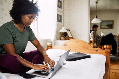 Young woman using laptop on bed while friend sitting in background at home