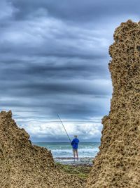 Man fishing on rock by sea against sky