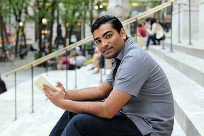 Portrait of man holding book while sitting on steps in city